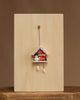 The Christian Ulbricht Ornament - Snowman with Red Cuckoo Clock, made in Germany, features a snowman and pine tree and hangs against a plain light wooden background on a centered wooden surface.