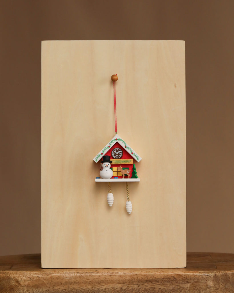The Christian Ulbricht Ornament - Snowman with Red Cuckoo Clock, made in Germany, features a snowman and pine tree and hangs against a plain light wooden background on a centered wooden surface.