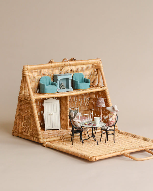 Miniature Braided A-Frame Dollhouse with a blue sofa set and fireplace inside, and two toy mice seated outside on a black wire table and chairs, on a soft beige background.