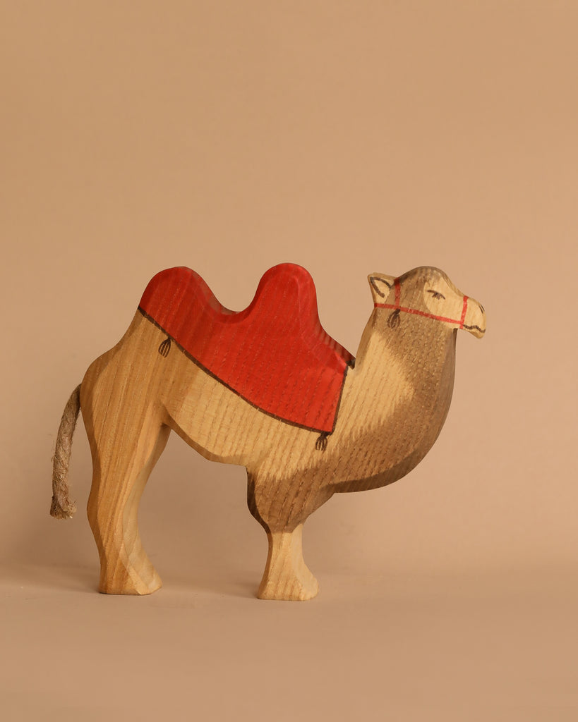 The Ostheimer Camel With Saddle is a beautifully handcrafted wooden figurine featuring a natural wood finish and a red, painted saddle. It stands proudly against a plain beige background, reminiscent of classic Ostheimer toys, and invites imaginative play.