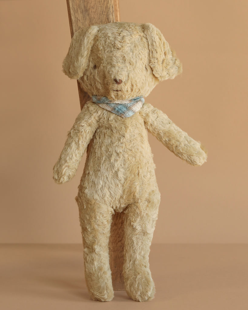 The Maileg Poodle Dog, Vintage Plush - Soft Sand stands upright with floppy ears and a blue and white checkered scarf. Made from cotton viscose plush, it’s displayed against a simple wooden plank and tan background.