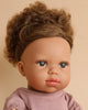 A close-up of a Minikane Baby Doll (13") - Melissa with curly brown hair tied up, large brown eyes, and freckles. The doll is wearing a light purple top and has a neutral facial expression. The background is a plain beige color.