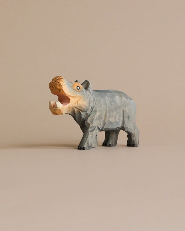 A Hand Carved Wooden Hippo toy, painted in shades of blue and gray, stands on a beige background with its mouth wide open, displaying carved teeth.