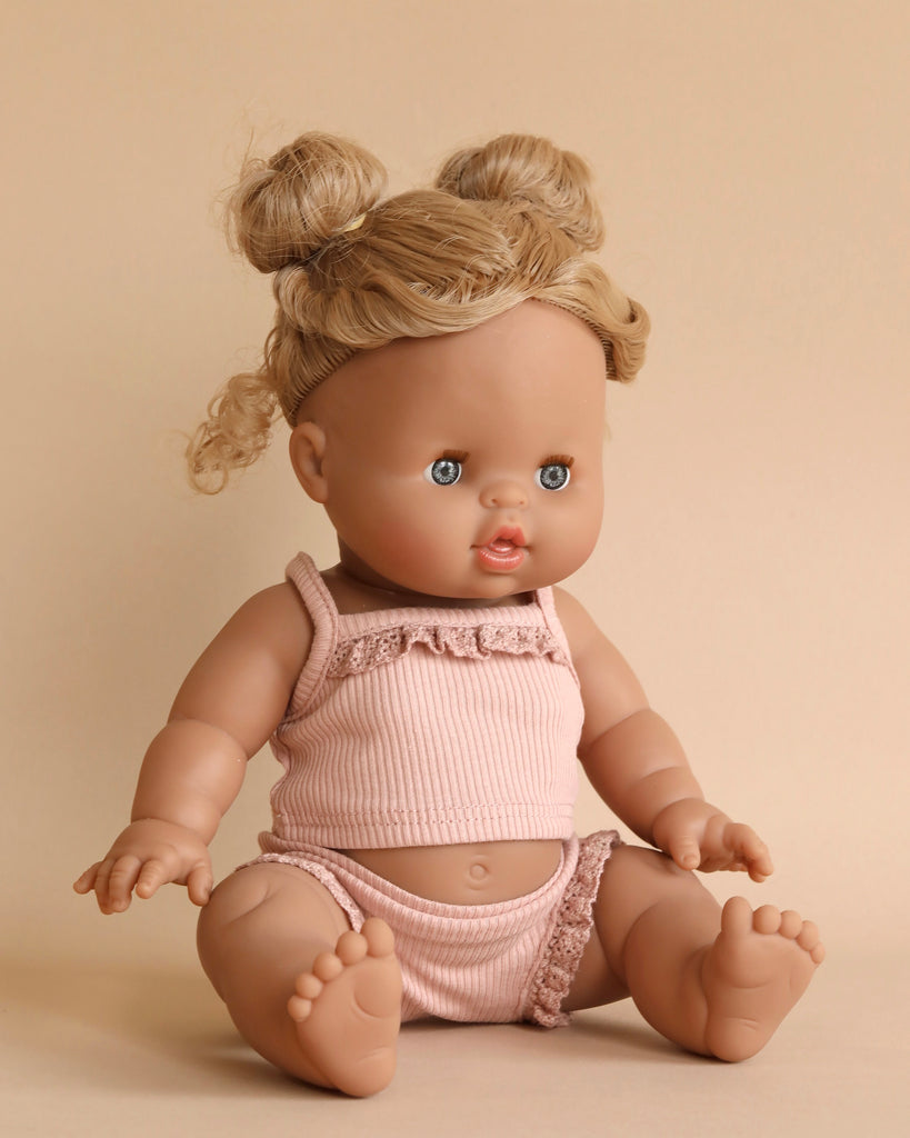 A Minikane Baby Doll (13") - Louise with light brown skin and blonde hair styled in two buns is sitting against a beige background. The doll, featuring blue eyes and an open mouth, is wearing a pink sleeveless top with a ruffled trim and matching bloomers. It includes anatomically correct details and a natural vanilla scent.