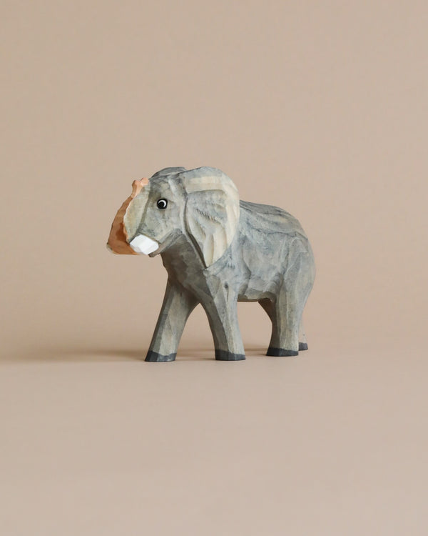 A Hand Carved Wooden Elephant figurine standing on a plain beige background. The elephant, intricately carved with visible grains and subtle color variations, serves as perfect kids' bedroom decor, emphasizing its trunk and large ears.