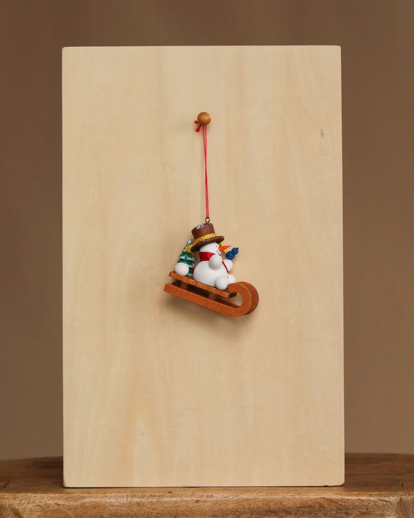 The Christian Ulbricht Ornament - Snowman on Sled features fine German craftsmanship, with a snowman in a hat and scarf on a sled, hanging from a red string against a wooden board on a simple background.
