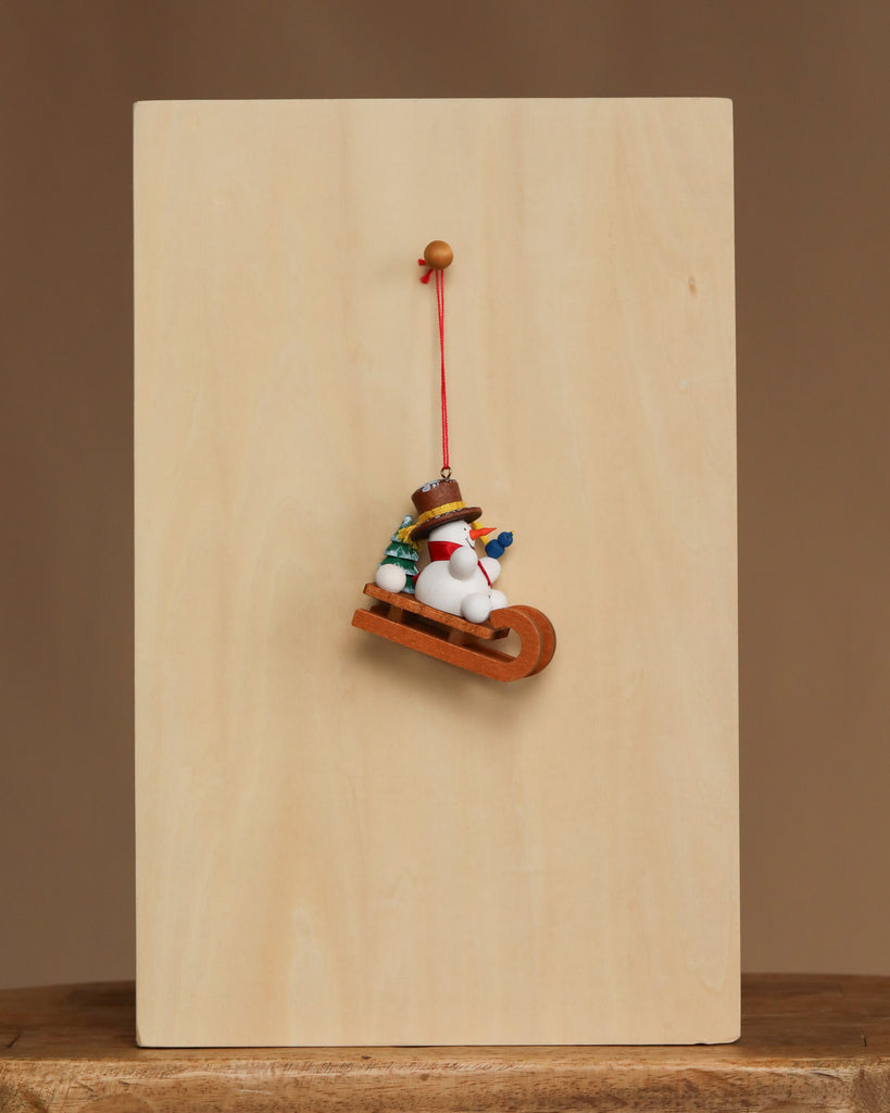 The Christian Ulbricht Ornament - Snowman on Sled features fine German craftsmanship, with a snowman in a hat and scarf on a sled, hanging from a red string against a wooden board on a simple background.