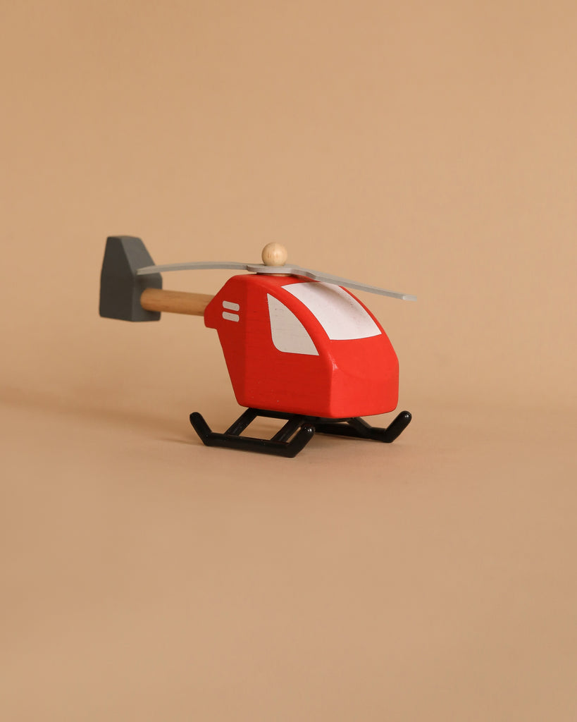 Introducing the "Helicopter," a delightful small wooden toy ideal for imaginative play. It boasts a striking red body, white windows, and gray rotors, all crafted sustainably. This charming helicopter stands out proudly against a simple beige backdrop.