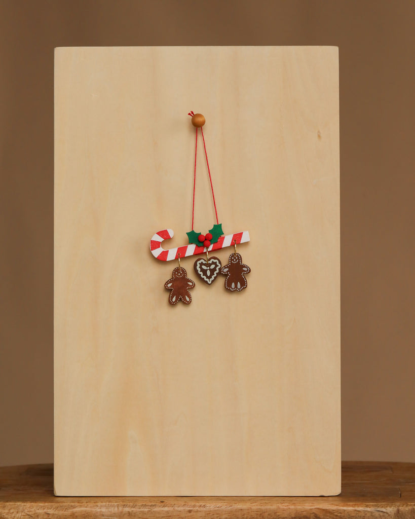 The Christian Ulbricht Ornament - Candy Cane Ornament, featuring red-and-white stripes with holly, hangs from a wooden peg beside a collectible plaque with two gingerbread people and a heart-shaped cookie on a plain wooden background.