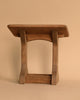 A small wooden stool, evoking the Ostheimer Nativity Scene, features a flat rectangular seat and two curved legs connected by a straight support beam, set against a plain beige background.