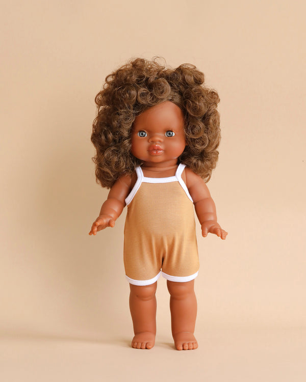 A Minikane Clothed Standing Doll (14.5") - Charlie with curly brown hair, brown eyes, and medium skin tone is dressed in a light brown romper with white straps and trim. The anatomically correct doll stands upright against a beige background and has a gentle vanilla scent.