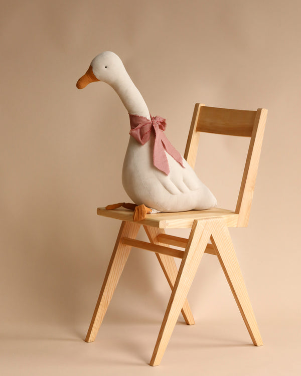 A Maileg Large Goose toy with a pink bow seated on a wooden chair against a beige background.