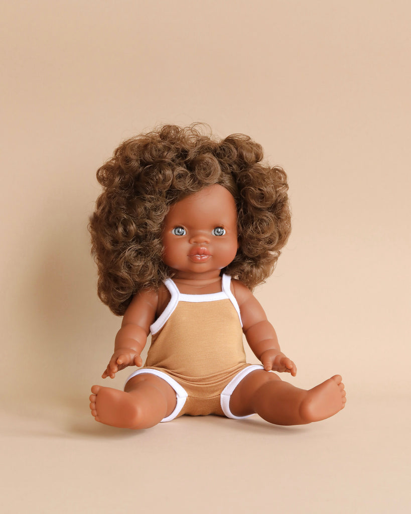 A Minikane Clothed Standing Doll (14.5") - Charlie is seated against a neutral beige background. The doll, which has a vanilla scent, is wearing a light brown romper with white trim and has a friendly expression with bright eyes. It is also anatomically correct for added realism.