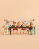 Six Maileg mice from the Maileg Family Celebration Set gather around a table filled with cake, fruits, veggies, milk, and honey. Set against a peach background, two mice sit comfortably in highchairs while four larger mice join them at the table—surrounded by whimsical Maileg accessories that complete this heartwarming scene.