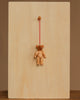 The Christian Ulbricht Ornament - Teddy Bear hangs by a red string from a round wooden peg on a plain, light wood board, creating a simple display with natural materials against a neutral brown background.