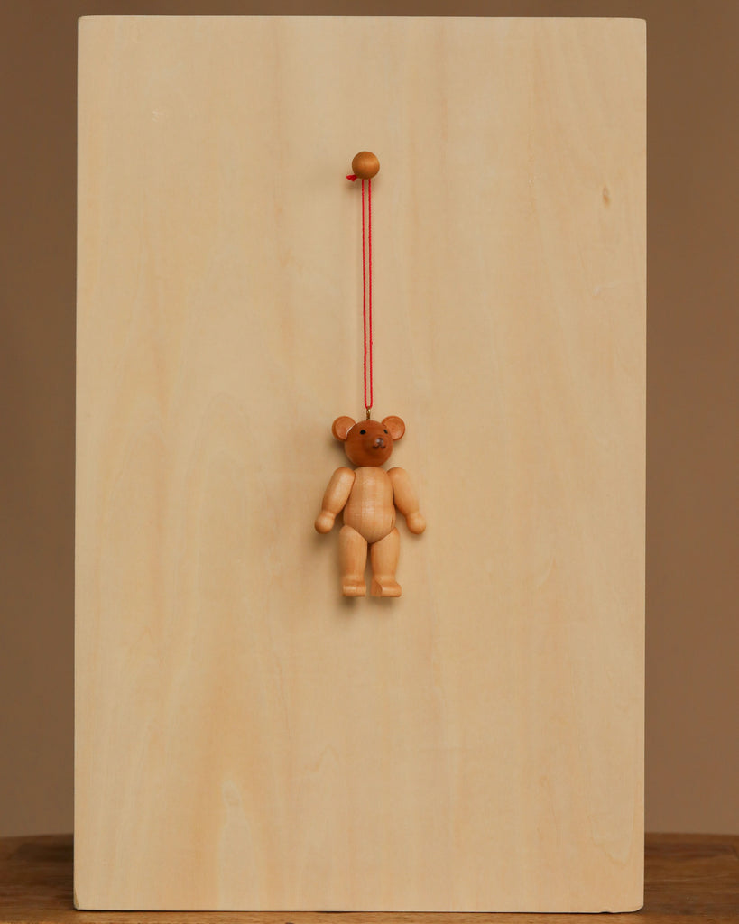 The Christian Ulbricht Ornament - Teddy Bear hangs by a red string from a round wooden peg on a plain, light wood board, creating a simple display with natural materials against a neutral brown background.