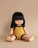 A Minikane Clothed Standing Doll (14.5") - Jade-Lou with straight black hair wearing a yellow romper with black trim is seated against a plain beige background, looking directly at the camera, exuding a soft vanilla scent.