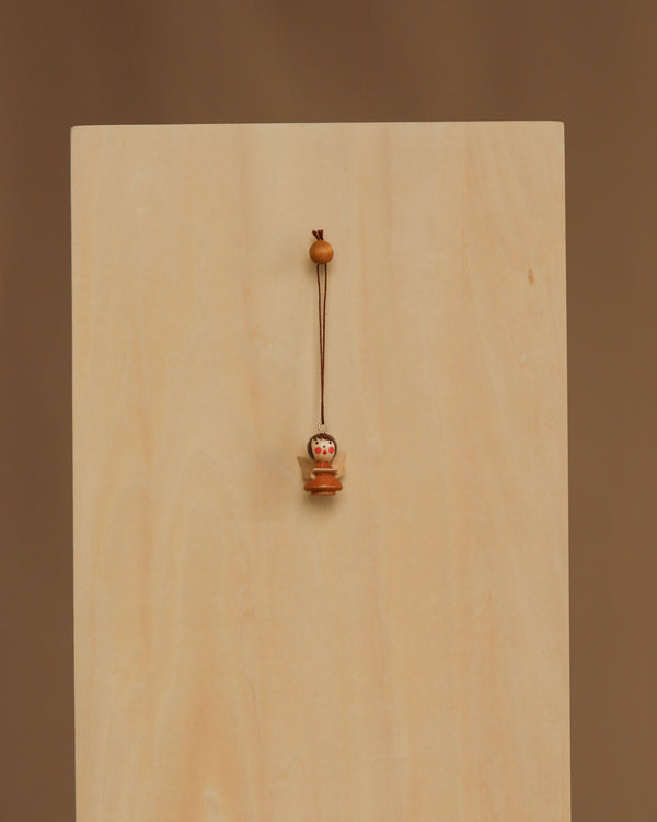The Christian Ulbricht Ornament - Angel (Natural) is a small wooden doll charm with a painted face, hanging by a string against a light wooden panel. Made in Germany, it contains small parts and may pose a choking hazard to young children.