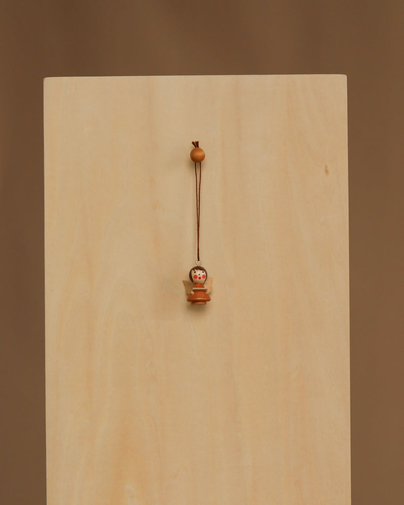 The Christian Ulbricht Ornament - Angel (Natural) is a small wooden doll charm with a painted face, hanging by a string against a light wooden panel. Made in Germany, it contains small parts and may pose a choking hazard to young children.