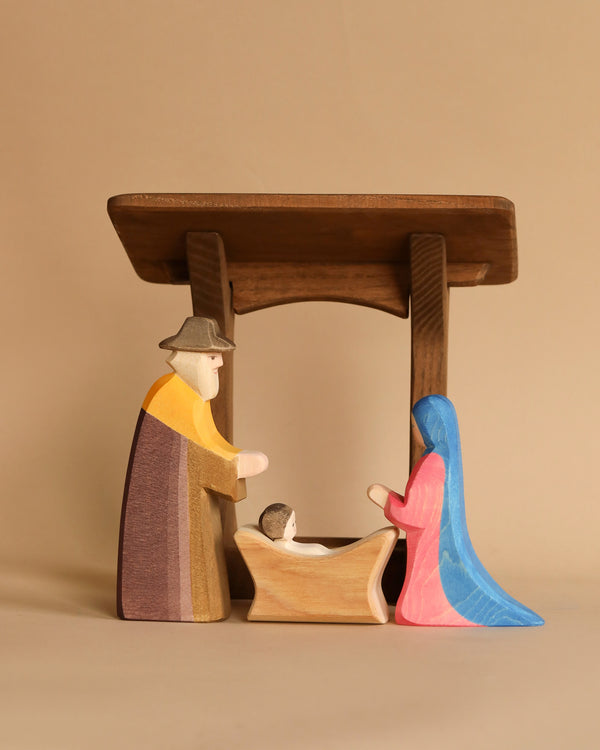 The Ostheimer Nativity Scene showcases a minimalist stable design that encourages imaginative play. It includes figures such as a character in a brown and yellow cloak, another dressed in a blue and pink robe, and an infant lying in a manger, all set against a gentle beige background.