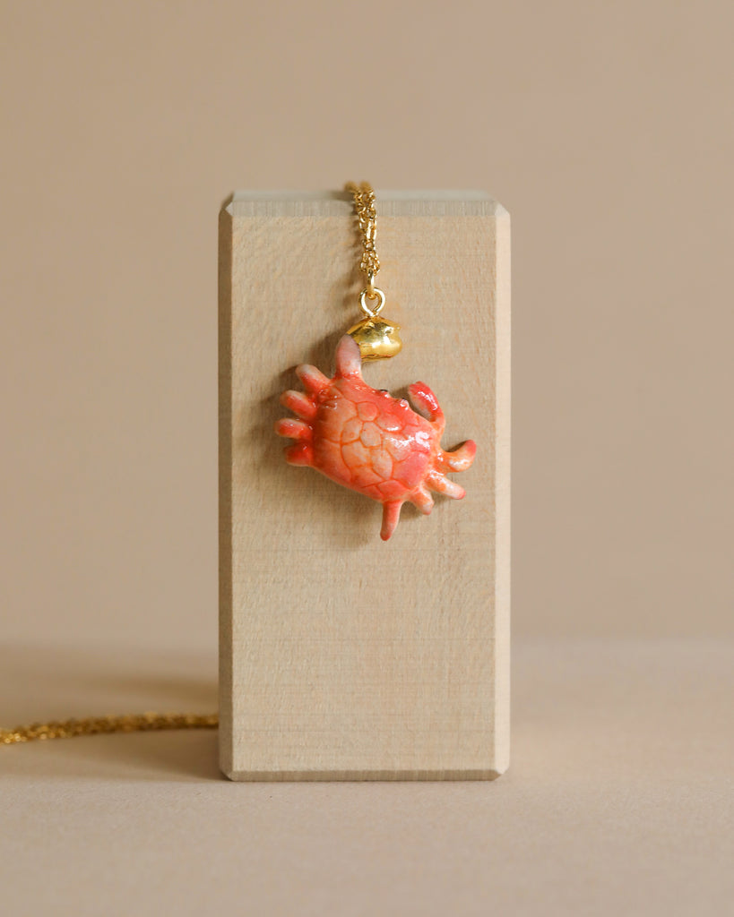 A delicate, anatomical heart pendant crafted from porcelain hangs from a Crab "Golden Grip" Necklace, displayed on a vertical, rectangular wooden block with a beige background.
