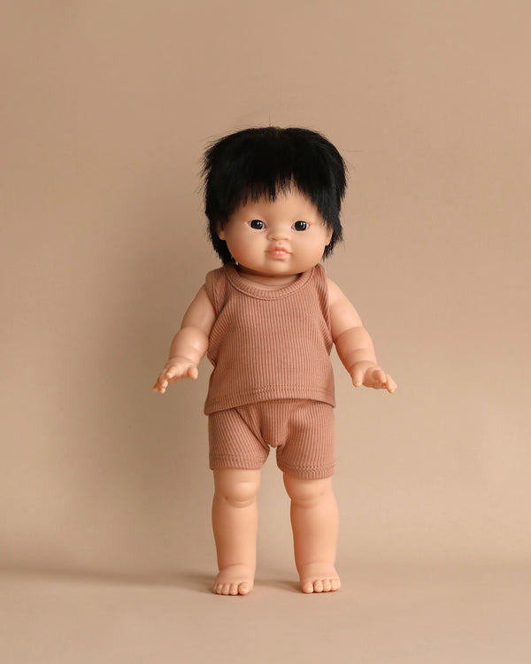 A Minikane Clothed Standing Doll (14.5") - Jude-Léo with straight dark hair and bangs stands against a beige background. The doll, featuring an anatomically correct design, is wearing a matching ribbed sleeveless top and shorts in a light brown color. Its arms and legs are slightly outstretched, exuding charm.