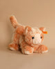 A Cassie Cat Plush Toy with orange and white fur sits on a neutral beige background. This huggable companion features a label attached to its ear, inviting you to embrace its soft charm.