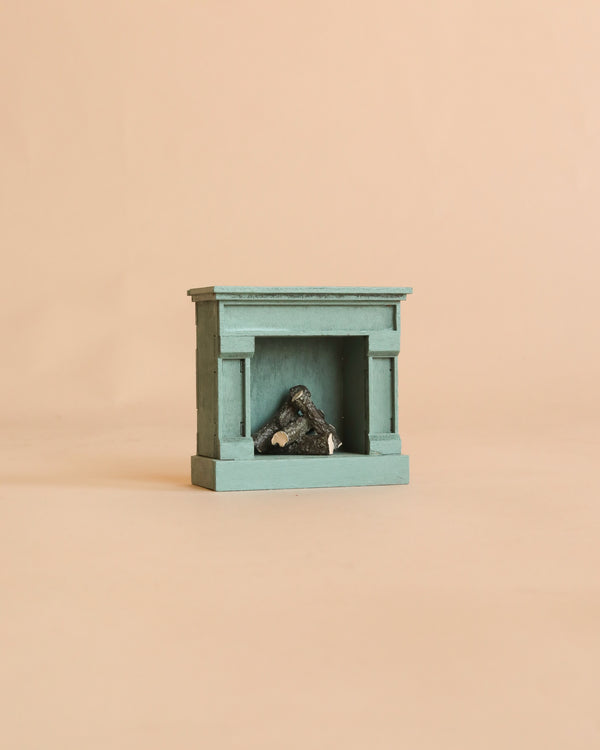 A Maileg Fireplace, Mouse—a small teal vintage fireplace featuring miniature logs inside—is displayed against a plain beige background.