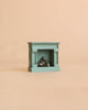 A Maileg Fireplace, Mouse—a small teal vintage fireplace featuring miniature logs inside—is displayed against a plain beige background.