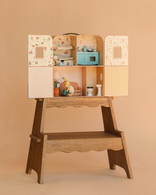 The Maileg Suitcasa Furnished Set (ships in one week) features pastel wallpaper and comes with miniature furniture and Mouse Size accessories, creating a cozy scene with a teddy bear reading in an armchair among potted plants and soft lighting.