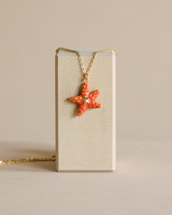 A coral-colored Starfish Necklace, hand painted and hanging on a delicate 24k gold plated steel chain, displayed against a vertical beige rectangular stand with a neutral background.
