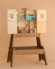 The Maileg Suitcasa Furnished Set features a compact dollhouse with opening doors, miniature furniture, kitchen, living space, and a small rabbit doll—all displayed on a wooden stand against a beige background. Ships in one week.
