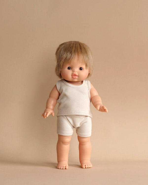 A Minikane Clothed Standing Doll (14.5") - Archie with straight blond hair and blue eyes stands against a beige background. The anatomically correct doll is wearing a sleeveless white ribbed shirt and matching shorts, carrying a subtle vanilla scent, and has an expressionless face.