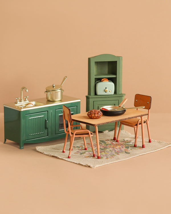 The Maileg Green Kitchen Set includes a green sink, stove, hutch, dining table with chairs, pots and pans, and a patterned rug, all displayed on a beige background.