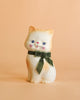 The Konges Sloejd Money Bank - Kitty is a handpainted ceramic cat figurine featuring blue eyes, light orange and white fur, and a green velvet bow around its neck, displayed against a plain peach background.