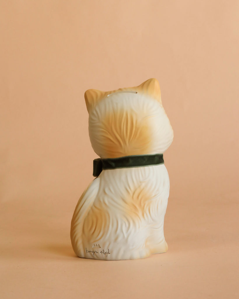 The Konges Sloejd Money Bank - Kitty is a handpainted ceramic cat figurine with cream and orange fur, shown sitting facing away and wearing a dark green collar, set against a solid peach-colored background.