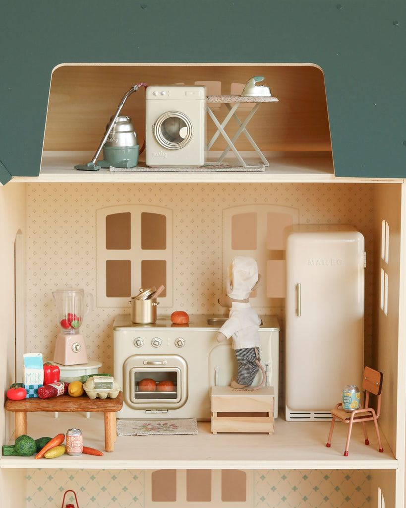 Create playful scenes with the Maileg Fully Furnished Dollhouse Set + Surprise Gift, featuring a toy chef, vintage stove, fridge, kitchen furniture, upstairs laundry area, and a peek at the miniature bathroom for added fun.