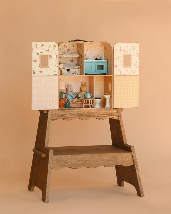 The Maileg Suitcasa Furnished Set sits on a wooden table, filled with dollhouse accessories and toy animals to create a charming scene, set against a plain beige background. Ships in one week.