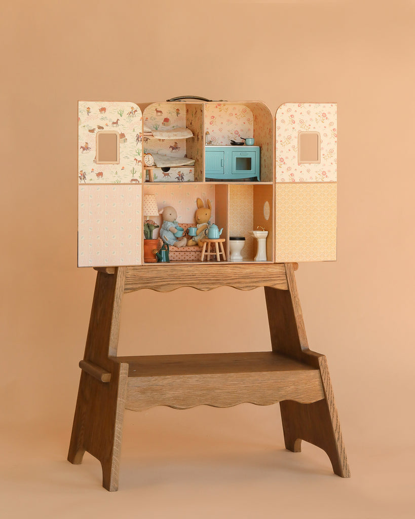 The Maileg Suitcasa Furnished Set sits on a wooden table, filled with dollhouse accessories and toy animals to create a charming scene, set against a plain beige background. Ships in one week.