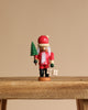 The Christian Ulbricht Collectible Wooden Nutcracker - Mini Santa stands on a wooden surface, holding a green tree and gift box. Dressed in red with a white beard and hat, it’s beautifully crafted against a plain beige background.