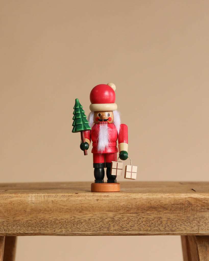 The Christian Ulbricht Collectible Wooden Nutcracker - Mini Santa stands on a wooden surface, holding a green tree and gift box. Dressed in red with a white beard and hat, it’s beautifully crafted against a plain beige background.
