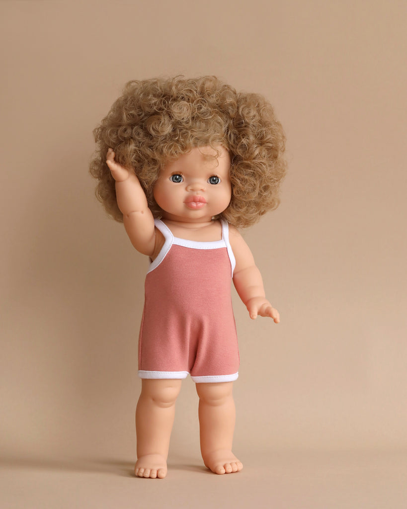 A Minikane Clothed Standing Doll (14.5") - Lise-Anaïs with curly, light brown hair stands on a neutral background. It is wearing a sleeveless pink romper with white trim. The anatomically correct doll has one arm raised and bare feet, and it is looking forward.