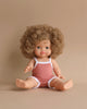 A cute, chubby-faced Minikane Clothed Standing Doll (14.5") - Lise-Anaïs with curly brown hair sits upright against a beige background. The doll is wearing a sleeveless, pinkish-red romper with white trim. One arm is extended forward, and both legs are stretched out, exuding a natural vanilla scent.