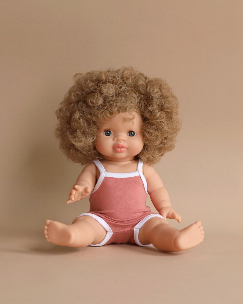 A cute, chubby-faced Minikane Clothed Standing Doll (14.5") - Lise-Anaïs with curly brown hair sits upright against a beige background. The doll is wearing a sleeveless, pinkish-red romper with white trim. One arm is extended forward, and both legs are stretched out, exuding a natural vanilla scent.
