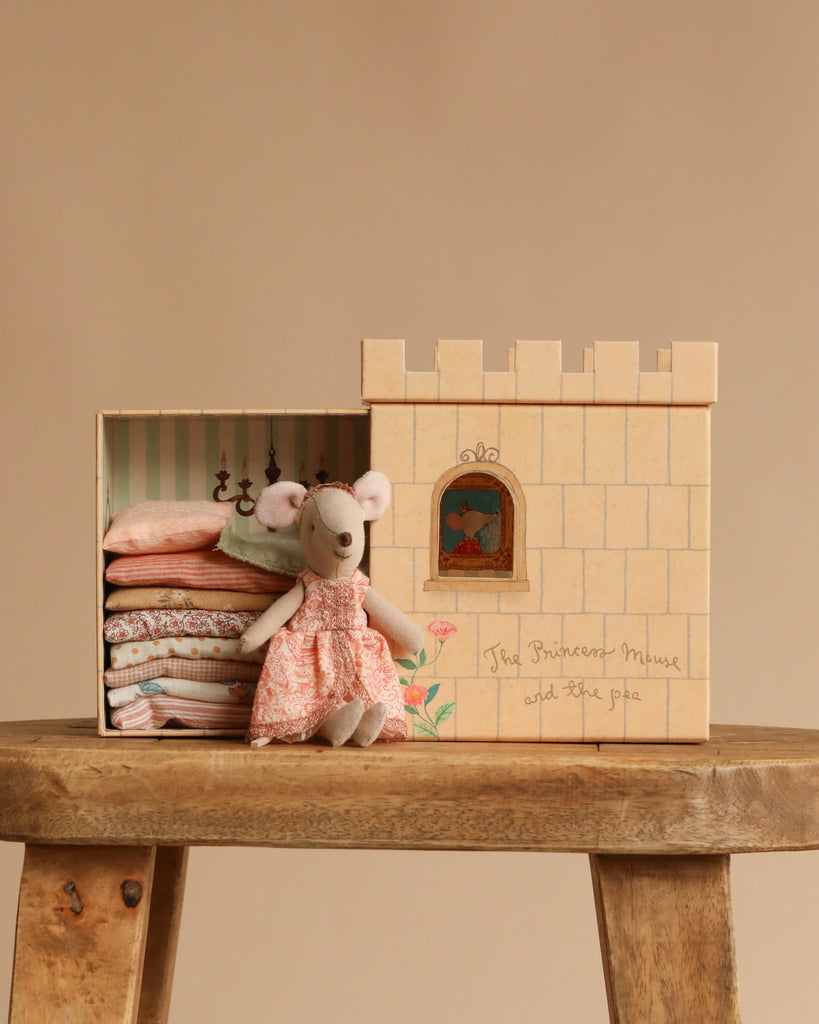 A delightful Maileg Fairytale Set features a small mouse in a pink dress sitting before a castle-shaped box filled with folded clothes, all arranged on a wooden stool, evoking the enchanting Princess and The Pea story.