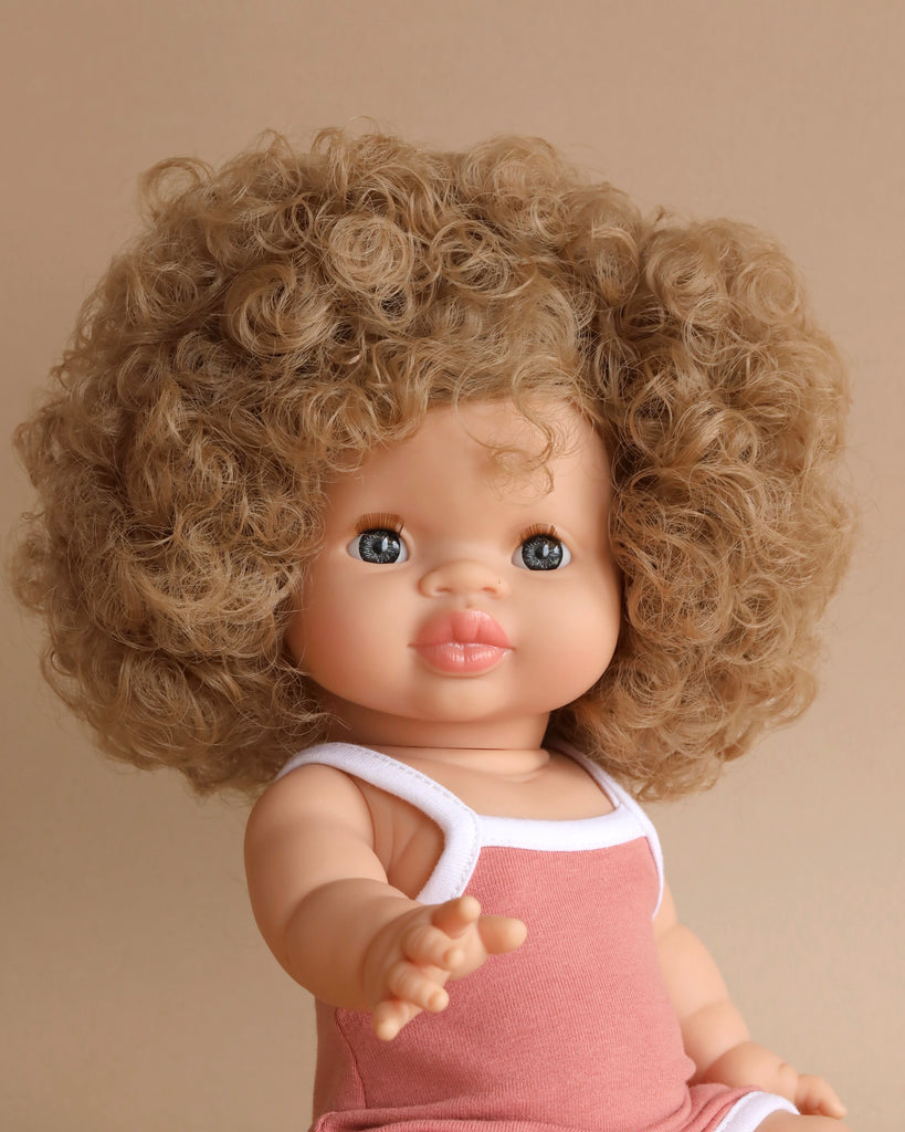 A Minikane Clothed Standing Doll (14.5") - Lise-Anaïs with big, curly blonde hair and light blue eyes is wearing a sleeveless pink outfit with white trim. The anatomically correct doll, known for its natural vanilla scent, stands against a neutral beige background with an outstretched arm.