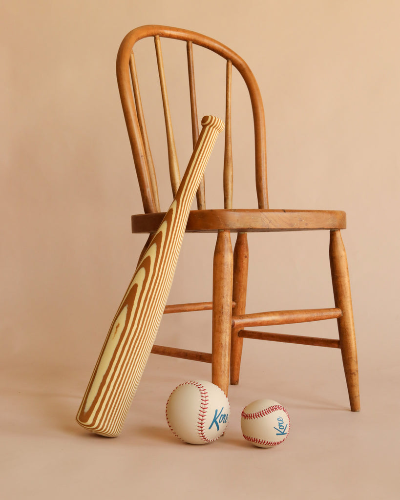 A Foam Baseball Bat + Baseball + Softball Set, arranged with the bat leaning against a wooden chair and two soft baseballs on the floor in front, all set against a beige background—perfect for safe indoor play.