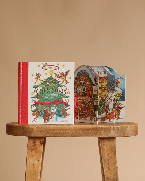 The children’s book "Christmas is Coming to Town!" with colorful Brown Bear Wood illustrations sits on a stool, beside a festive pop-up scene featuring animals, a house, Christmas tree, and holiday decorations against a beige background.
