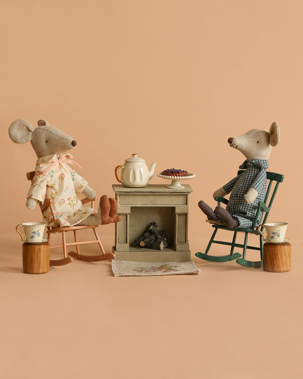 The Maileg Grandma and Grandpa Afternoon Tea Set features two mice in rocking chairs by a small table with a miniature tea set and dessert, creating a cozy scene with a vintage fireplace and neutral beige background—perfect for tiny gatherings.