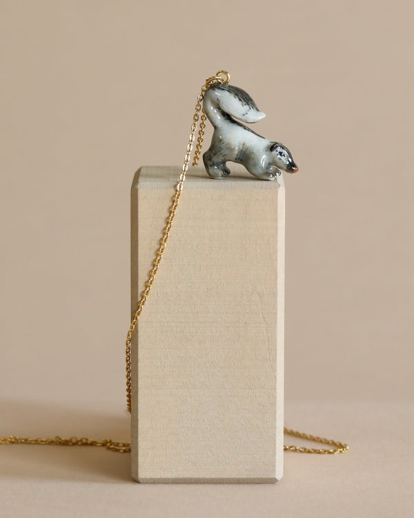 A pendant necklace featuring a silver skunk, suspended on a thin 24k gold plated steel chain, draped over a tall beige box against a neutral background.